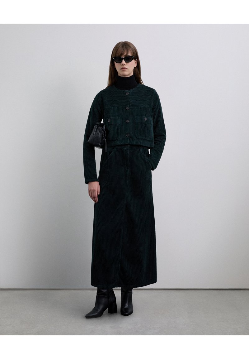 Veste courte en velours côtelé vert foncé avec deux poches avant, associée à une jupe longue assortie. Le modèle porte des lunettes de soleil noires et des bottines.