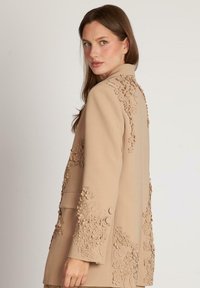 Sacou bej cu embellismente florale pe mâne și spate, având un design structurat, guler șanț și accente cu butoane pentru închidere.