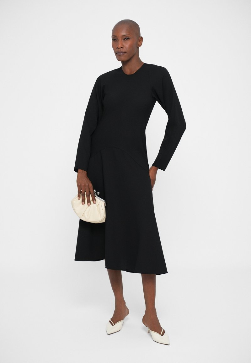 Femme en robe midi noire à manches longues tenant une petite pochette beige, portant des escarpins à talons kitten blancs, se tenant devant un fond blanc.