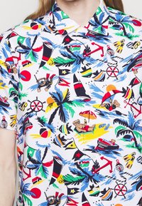 Camisa de manga corta con un estampado tropical colorido que incluye palmeras, escenas de playa, barcos y animales sobre un fondo blanco.