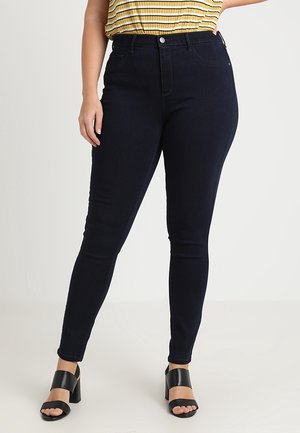 Mujer con vaqueros pitillo de cintura alta de color azul oscuro, top amarillo con rayas metido dentro y sandalias negras de tacón, de pie frente a un fondo liso.
