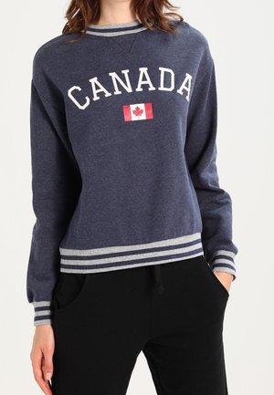 Person iført en mørkeblå sweatshirt med "CANADA" og et canadisk flag, kombineret med sorte bukser, hænderne i lommerne.