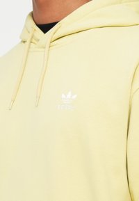 Oseba nosi svetlo rumeno adidas kapuco z belim trefoil logotipom in vrvico čez črno majico z dolgimi rokavi, razkrivajoč vrat in ramena.