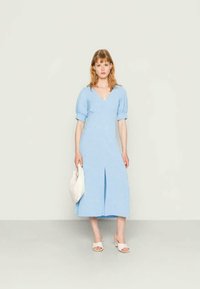 Mulher numa vestido midi azul claro com mangas bufantes, segurando uma mala branca, usando sandálias de salto brancas, a pousar contra um fundo simples.