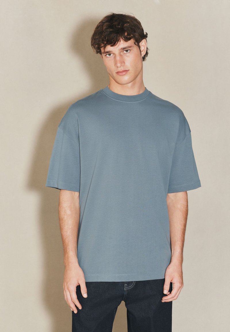 Tricou supradimensionat albastru deschis din material moale, cu gât rotund și mâneci scurte. Asortat cu blugi închiși la culoare, pe un fundal neutru.