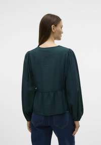 Mørkegrøn, tekstureret bluse med scoop halsudskæring, puffede lange ærmer og samlet talje. Sammen med højtaljede blå jeans.