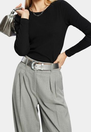 Vrouw draagt een zwart shirt met lange mouwen, grijze, geplooide broek met hoge taille en een zilveren riem, en houdt een zilveren handtas over haar schouder.