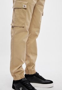 Beige cargobroek met elastische boorden en zijzakken, gecombineerd met zwarte sneakers met een witte zool en logo-accent.