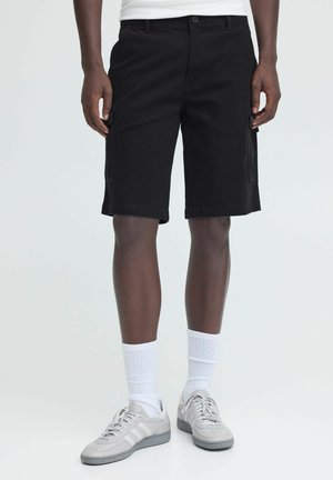Man draagt zwarte cargoshorts tot op de knie, witte sokken en lichtgrijze sneakers, staand tegen een effen achtergrond.