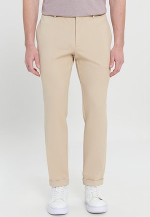 Mand iført beige slim-fit bukser med opkørte manchetter og hvide sneakers, stående mod en ensfarvet lys baggrund.