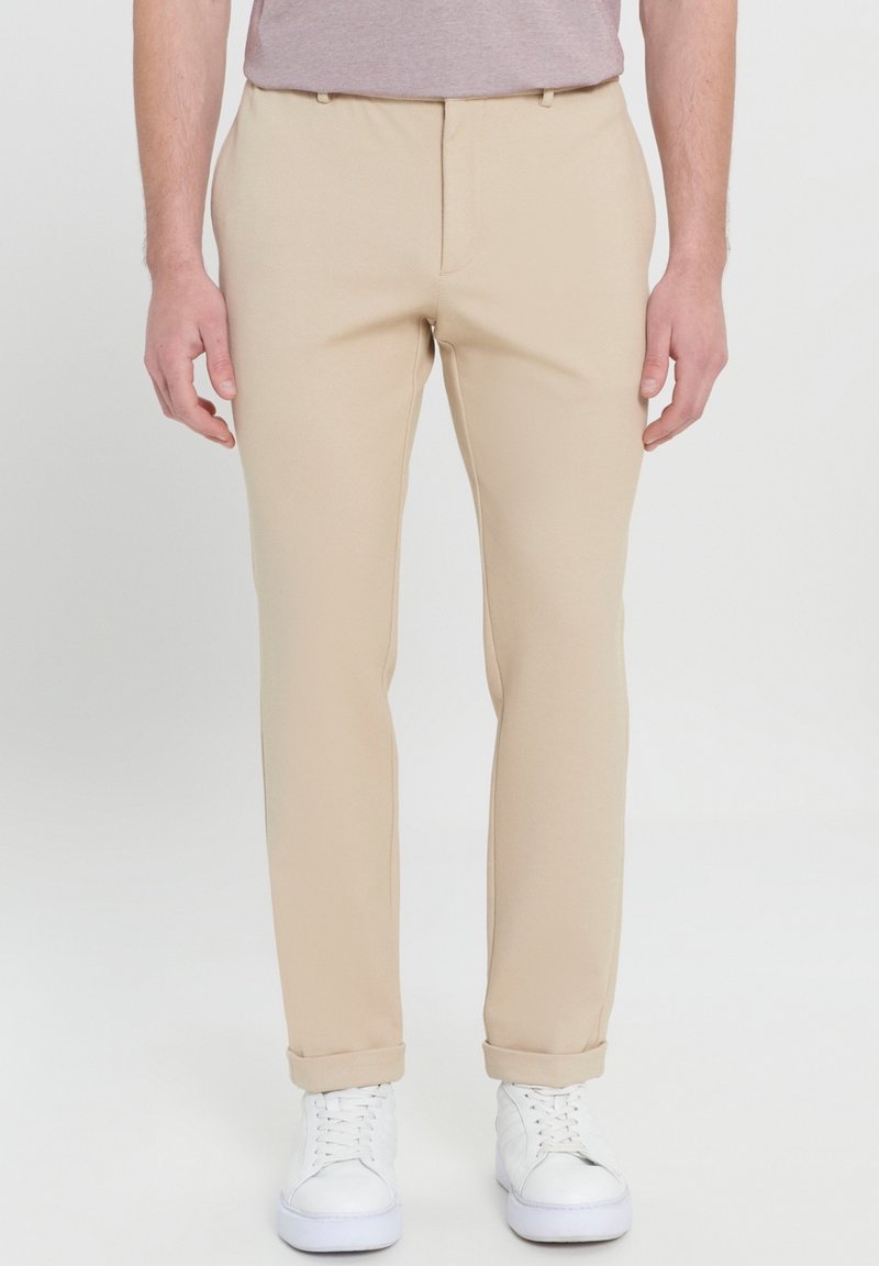 Homme portant un pantalon beige slim fit avec les revers roulés et des baskets blanches, debout devant un fond clair uni.