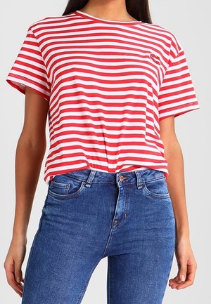 Vrouw draagt een rood-wit gestreept T-shirt met korte mouwen, ingestopt in een hooggesneden blauwe spijkerbroek met zichtbare zakken en knoop.