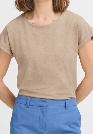 Kvinde iført en beige T-shirt med korte ærmer stoppet ned i højttaljede blå bukser med bæltestropper, stående med hænderne bag ryggen.