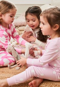 Kolm last, kes kannavad pidžaamasid roosade südamete ja täppidega, hoiavad pehmeid kaisuküülikuid. Taustal on tekstuuriga naturaalsetest kiududest matt.