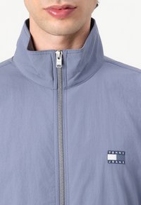 Homme portant une veste zippée bleu clair à col montant avec le logo Tommy Jeans sur la poitrine, gros plan sur le cou et le haut du torse.
