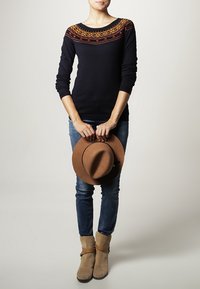 Marineblauer Pullover mit mehrfarbigem geometrischem Strickdesign am Kragen, kombiniert mit blauen Jeans, beige Ankle Boots und einem braunen Hut.