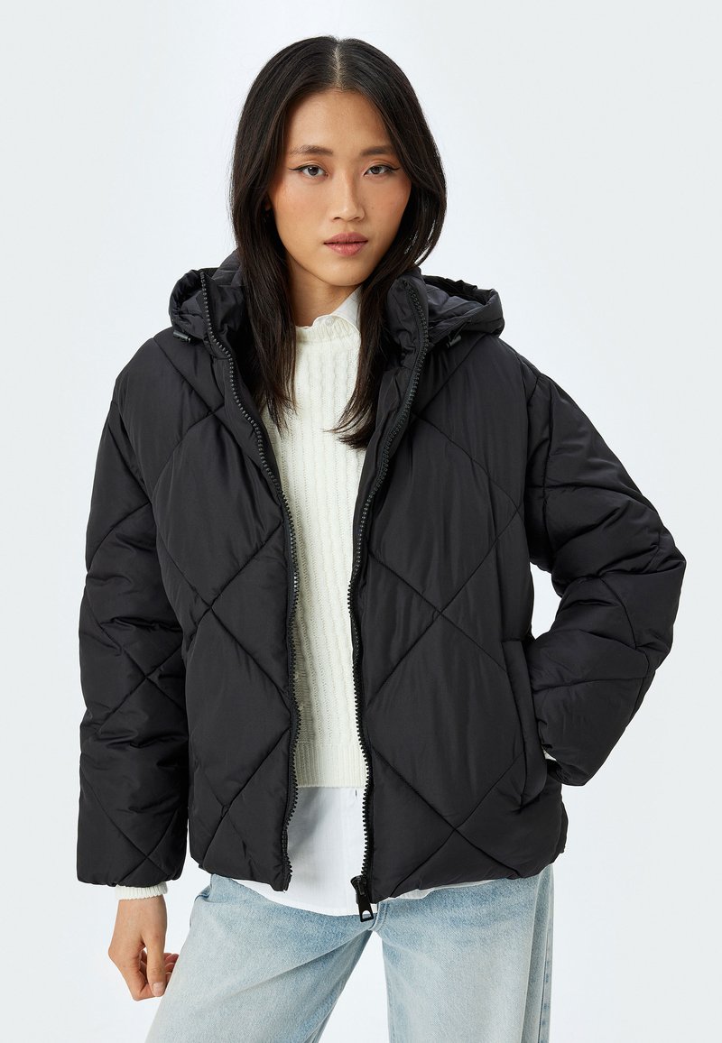 Sort quiltet pufferjakke med hætte, med en frontlynlås og diamantmønster. Båret over en hvid strikket bluse og lyse jeans.
