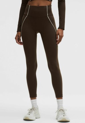 Personne portant des leggings taille haute marron foncé avec un liseré blanc, des chaussettes blanches et des chaussures de sport blanches, debout devant un fond uni.