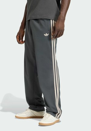 Mann trägt dunkelgraue Adidas-Trainingshose mit beigen Streifen und Logo, kombiniert mit beigen Sneakers mit Gummisohlen, stehend auf hellem Hintergrund.