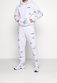 Grauer Pullover mit Schmetterlingsmustern, Kapuze und Vordertasche. Kombiniert mit passenden Jogginghosen mit demselben Schmetterlingsdesign und Logo.