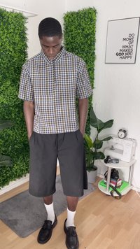 Jaunas vyras apsirengęs languota trumpomis rankovėmis marškinių ir juodais šortais stovi patalpoje šalia žalių augalų ir stalo su kamera, knygomis bei siuvimo mašina.