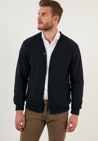 Tums tumši jūras spēku kardigans, kas izgatavots no adīta auduma, ar V veida kaklu, pogu priekšpusi un ribotām aprocēm. Uzvilkts pāri baltai kreklam.