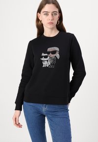 Svart sweatshirt med en grafik av två karaktärer med solglasögon, prydd med ett glittrande rhinestonesmönster. Långa ärmar och rund halsringning.