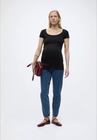 Terhes nő áll, fekete testhezálló pólót, kék farmert, piros cipőt visel, és bal kezében piros kézitáskát tart.