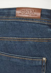 Mørkeblå denimjeans har et brunt lærmerke med "ONLY" preget, kontrasterende gult søm, og en metalknappdetalj ved lommen.
