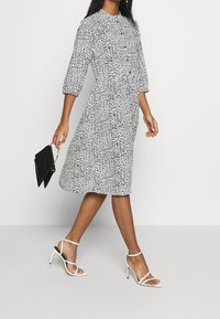 Abito a righe in bianco e nero con colletto, maniche tre quarti e vita stretta, abbinato a tacchi bianchi con strappi e una borsa nera.