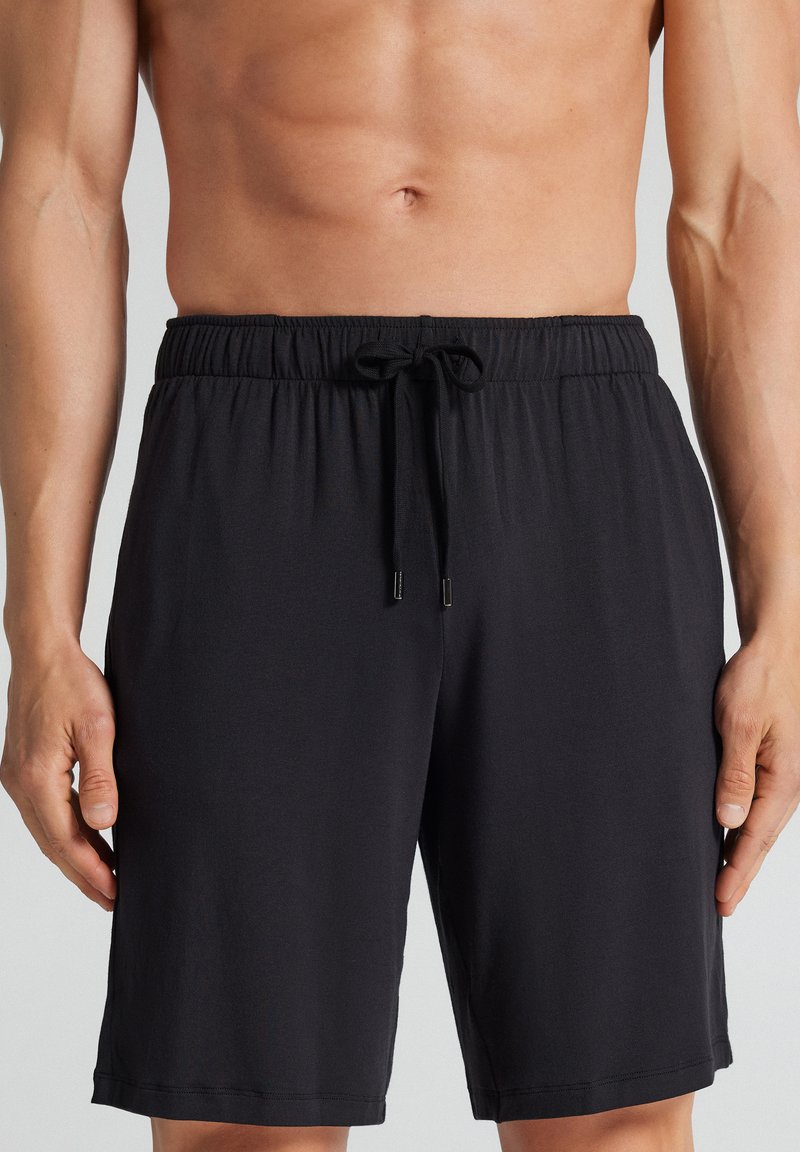 Männlicher Torso mit einfachen schwarzen Shorts mit Kordelzug, der eine nackte Brust und Arme vor neutralem Hintergrund zeigt.