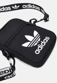 Černá křížová taška vyrobená z textilie se zipovou kapsou, která má výrazné bílé logo Adidas a nastavitelný popruh s logem.
