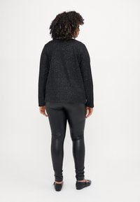 Must mustaong varrukatega tipp, millel on tekstuuriga metalliline muster, koos mustade kunstnahast legging'itega ja mustade tasaste kingadega, vaadates tagant.