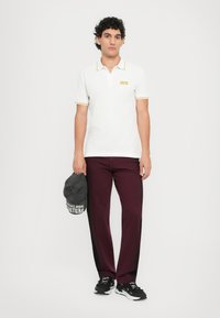 Chemise polo blanche avec des finitions jaunes, pantalon bordeaux et baskets noires. Tient une casquette grise avec un texte blanc. Design décontracté et ajusté.