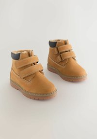 Tanfarbene Wildleder-Stiefeletten verfügen über zwei verstellbare Klettverschlüsse, ein Futter aus Kunstfell und eine Gummisohle mit einer gesteppten Nahtdetails.