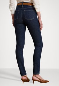 Donkerblauwe skinny jeans met een middenhoge pasvorm, voorzien van twee achterzakken, denimtextuur en een bruine riem. Gestyled met bruine hakken.