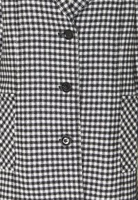 Sort-hvid ternet frakke lavet af tekstureret stof, med seks sorte knapper, to sidelommer og et klassisk kravedesign.