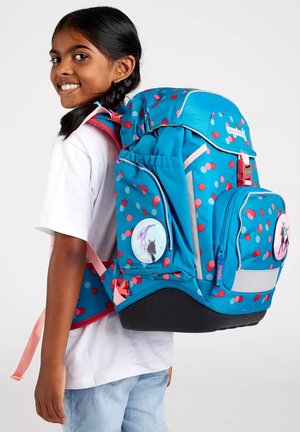 Smiling girl with braided hair wearing a large blue backpack with red and pink dots and unicorn patches, dressed in white shirt and jeans.