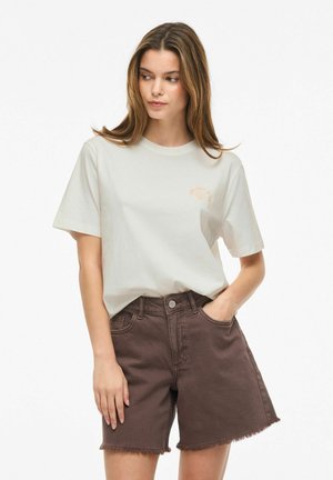 Joven mujer con cabello castaño claro que lleva una camiseta lisa de color blanco roto y pantalones cortos vaqueros marrones desgastados de talle alto, con una mano en el bolsillo.