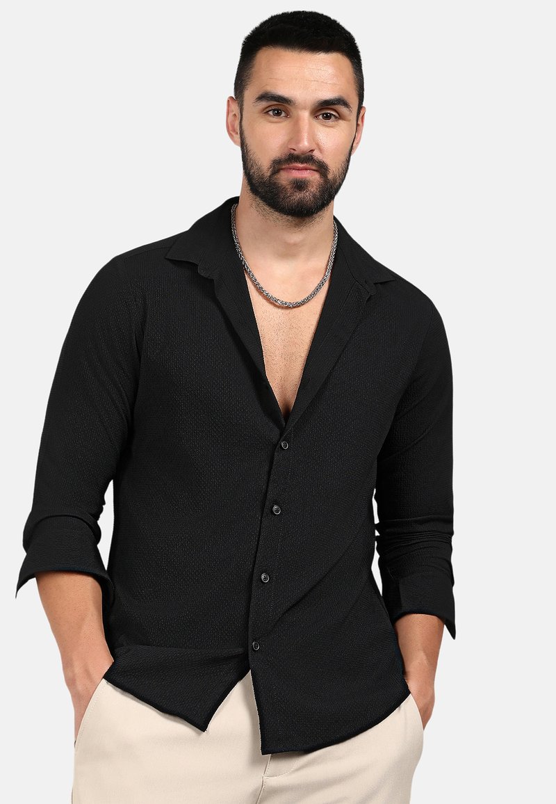 Camicia nera a maniche lunghe con bottone, pattern testurizzato, colletto aperto e design aderente. Bottoni frontali e maniche arrotolate visibili.