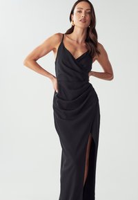 Schwarzes Abendkleid aus glattem Stoff, mit V-Ausschnitt, Drapierung und einem hohen Schlitz an einer Seite. Dünne Träger runden den Look ab.