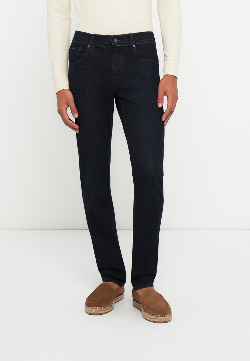 Tumed sinised denim püksid sirge jala disainiga, viie taskuga stiil ja nööbiga kinnitamine, koos heledast pruunist nahast libisevate kingadega.