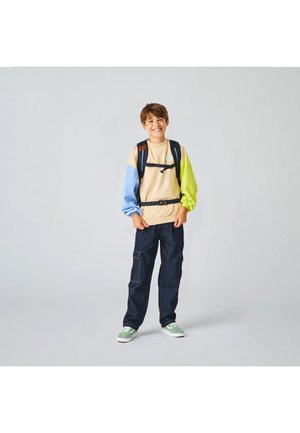 Ragazzo sorridente che indossa una felpa beige con maniche blu e verdi, pantaloni cargo scuri, scarpe da ginnastica verdi e uno zaino nero, su sfondo uniforme.