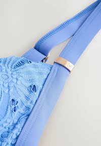 Blauer Stoffbeutel mit strukturierter Stickerei und Ausschnitt-Details. Verfügt über einen glatten blauen Riemen und ein roségoldfarbenes Metallelement nahe der Befestigung.