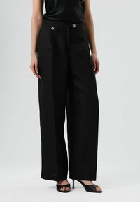 Zwarte broek met wijde pijpen van een gestructureerde stof, voorzien van voorste plooien, twee knopen in de taille en een losse, vloeiende pasvorm.