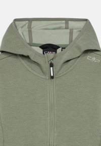 Πράσινη υφασμάτινη ζακέτα με φερμουάρ και κουκούλα, διαθέτει λείο εσωτερικό γέμισμα και μικρό λογότυπο κοντά στο γιακά. Ο φερμουάρ είναι μαύρος με ασημένιες λεπτομέρειες.