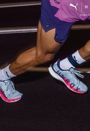 Läufer mit weißen Puma-Socken und blauen Puma-Laufschuhen mit roten Akzenten auf der Laufbahn, sichtbaren muskulösen Beinen und violetten Shorts.
