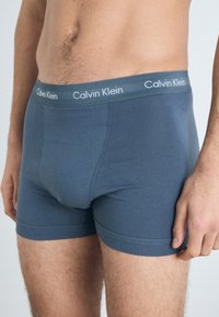 Dark blue boxer briefs made of a soft fabric, featuring a contrasting gray Calvin Klein logo waistband, with a snug fit and smooth texture.