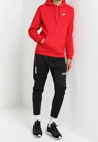 Rode Nike hoodie met kangoeroezak en logo, gecombineerd met zwarte joggerbroeken met zijstrepen en witte tekst. Zwarte sportschoenen.