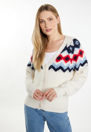 Junge Frau mit langen blonden Haaren, die eine weiße gemusterte Strickjacke und blaue Jeans vor einem schlichten hellen Hintergrund trägt.